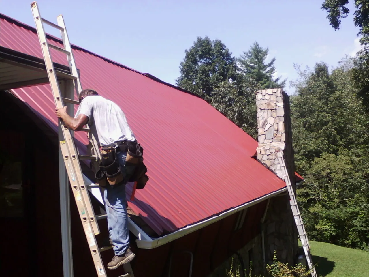 Roofer on a ladder beside a red metal roof and stone chimney — Soffit & Fascia Repair by Roofing Brothers in Reedsville